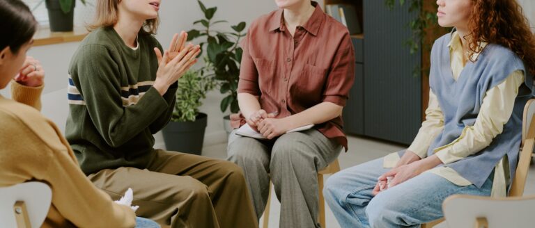 Group therapy session with four individuals discussing mental health issues. Participants sharing personal experiences in supportive environment with plants and clock in background