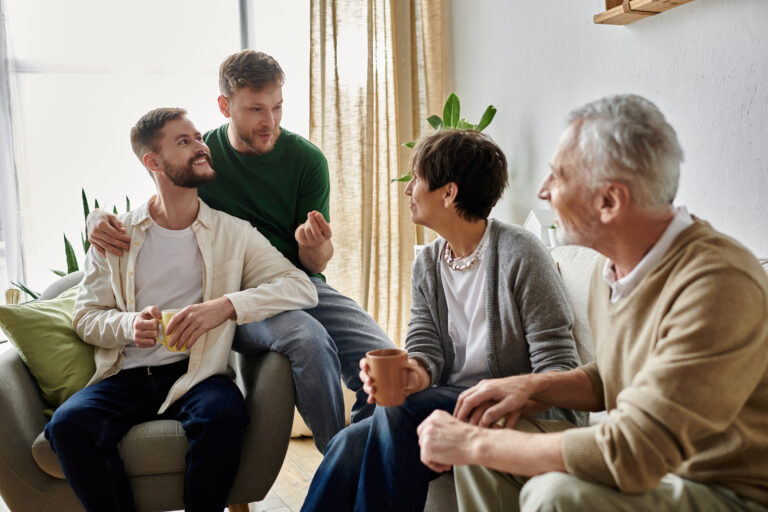 A gay couple sits with parents in their living room, enjoying a conversation and drinks.